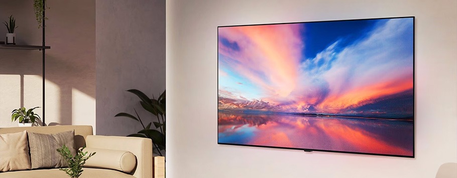 A modern living room with a wall-mounted flat-screen TV displaying a vibrant sunset over water. A beige sofa with pillows and potted plants adds warmth.