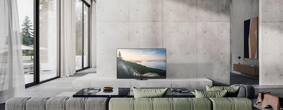 A modern, minimalist living room with large windows showing a forest view. A green sofa faces a flat-screen TV on a concrete wall. The space feels serene and airy.