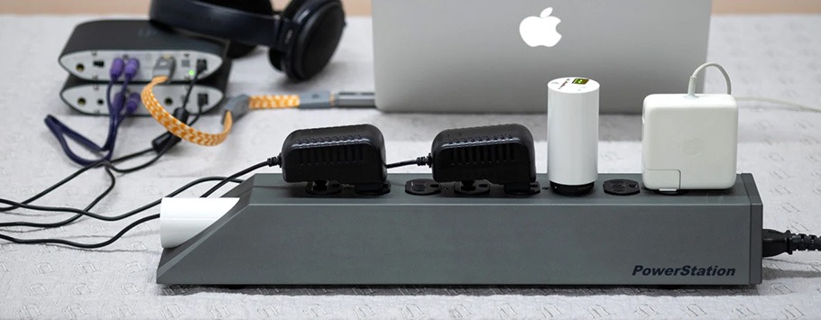 Power strip labelled "PowerStation" with multiple chargers on a textured table. Background includes a laptop, monitor, headphones, and cables.