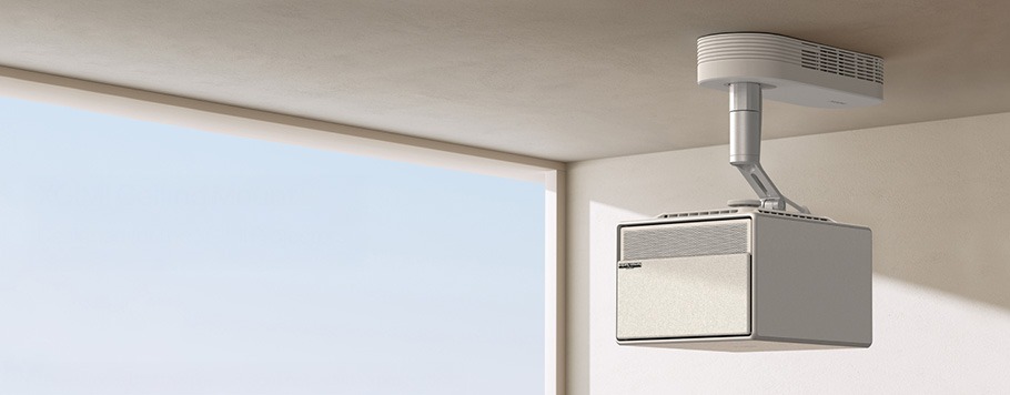 Ceiling-mounted modern projector against a plain, light-coloured wall with blue sky visible through a window. Clean, minimalist design with a sleek, futuristic feel.