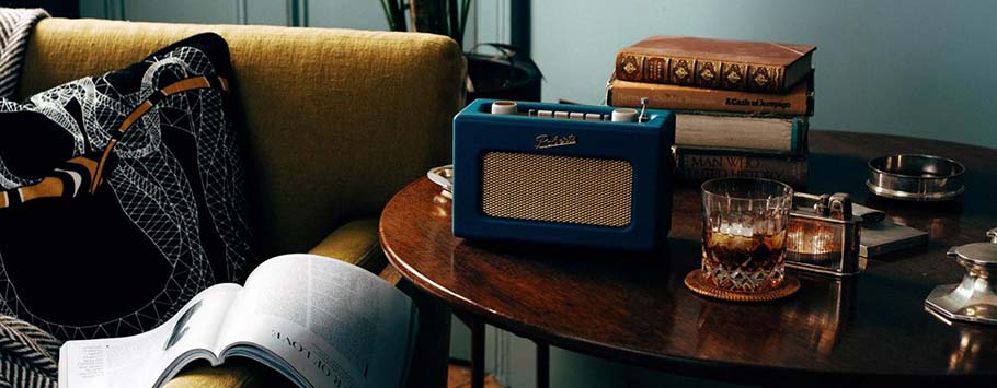 A cosy vintage scene with a blue retro radio on a wooden table, surrounded by stacked books and a glass of whiskey. Nearby, an open book and a patterned cushion rest on a mustard sofa.
