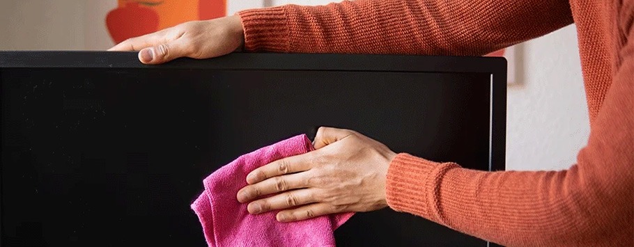 Hands in a rust-coloured sweater gently clean a TV screen with a pink cloth, conveying tidiness and care in a domestic setting.