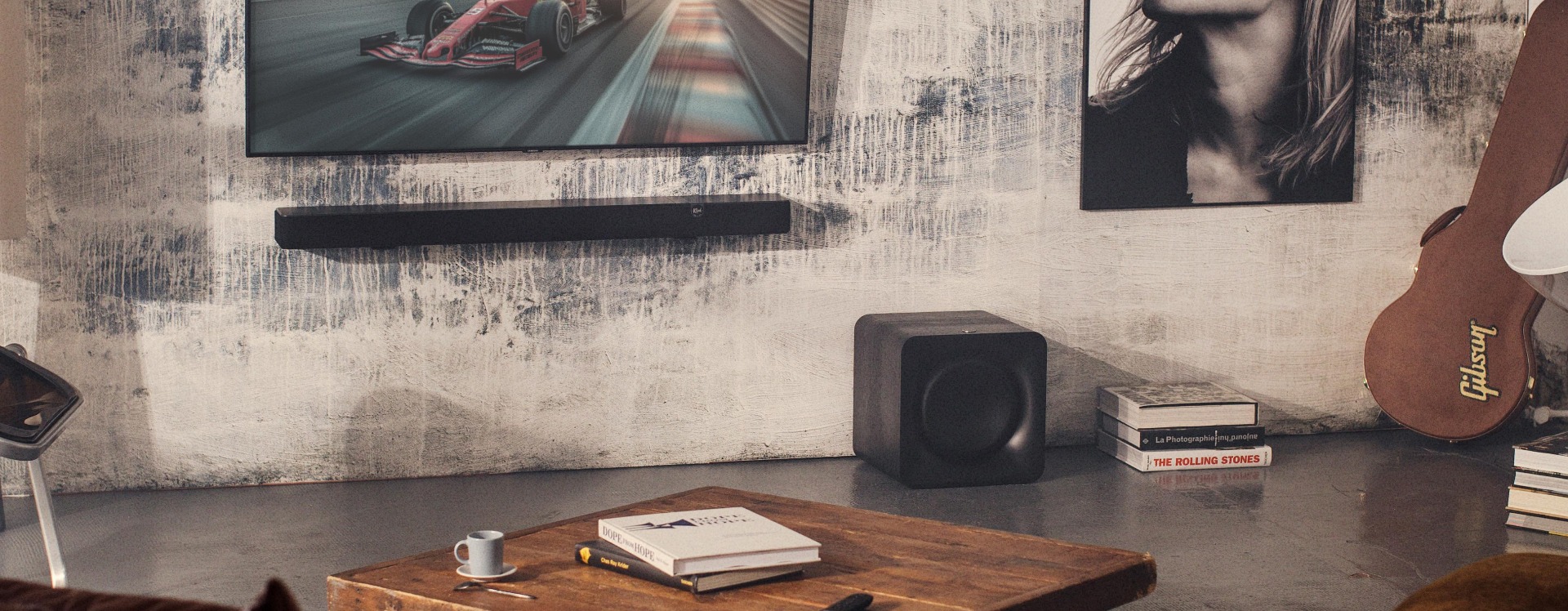 Living room with a sleek wall-mounted TV showing a race car, a soundbar below, a wooden table with books and coffee, and a guitar case nearby.