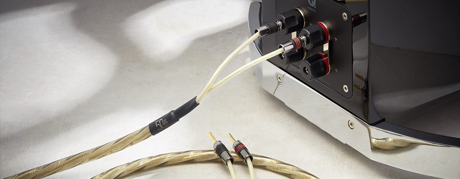 Close-up of premium audio cables connected to a sleek black speaker. The cables have transparent sheaths and gold connectors, conveying a high-end tech vibe.