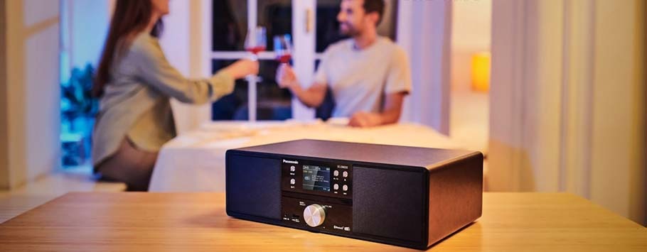 A modern stereo system is positioned on a wooden table, with a man and woman in the background toasting wine glasses.