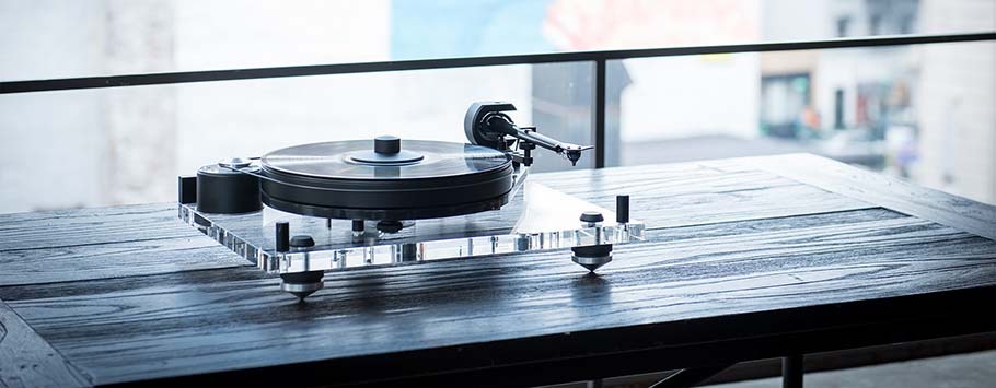 A sleek turntable with a transparent acrylic base sits on a wooden table, set against a blurred cityscape background, evoking a modern and nostalgic ambiance.