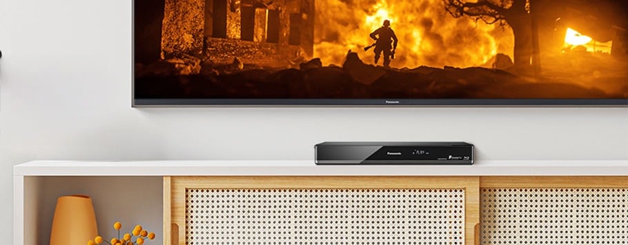 A TV displays a dramatic scene with a person silhouetted by orange flames. Below, a sleek media player sits on a white cabinet with a decorative vase.