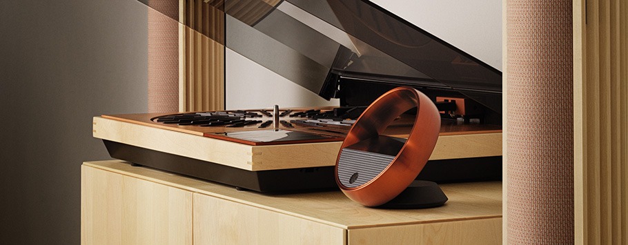 A sleek, modern turntable with a clear lid sits on a light wood cabinet. Nearby, a unique circular tv remote in copper adds a stylish, contemporary touch.