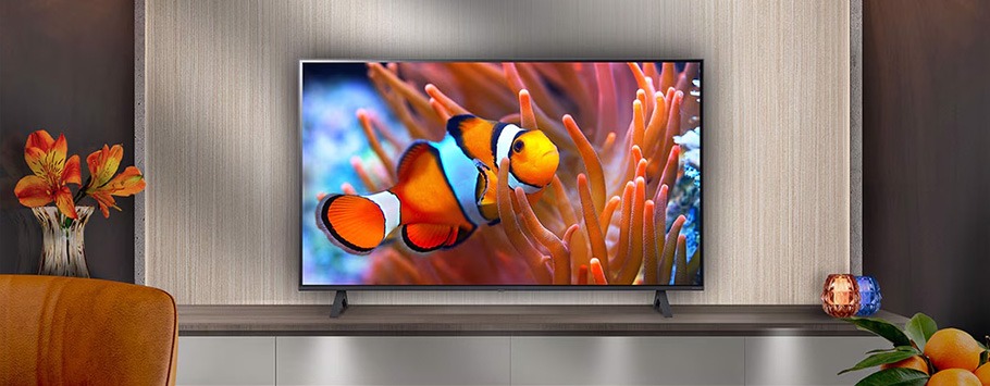 A vibrant clownfish swims among orange coral on a large TV screen centred on a modern wooden cabinet. A vase with orange flowers sits to the side.